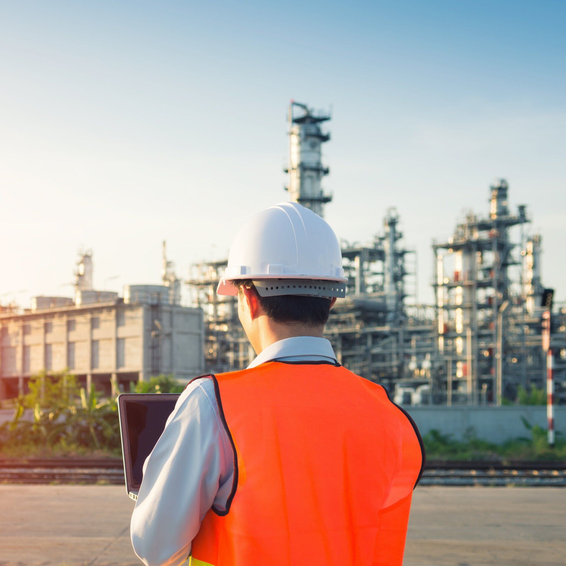 A man wearing a hard hat and an orange vest is standing in front of an oil refinery.