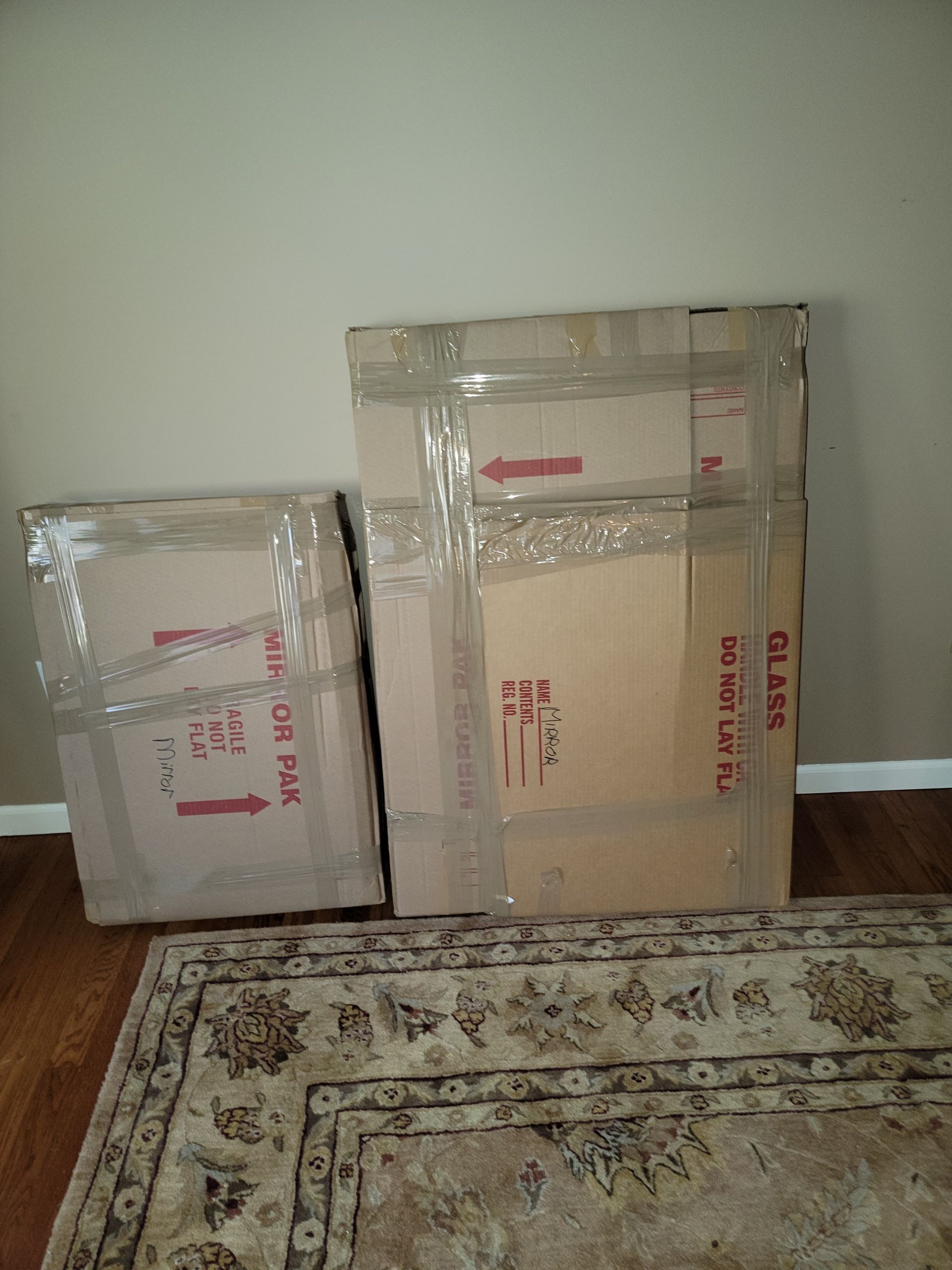 Two cardboard boxes, one large, taped together, against a wall, on a patterned rug, hardwood floor.