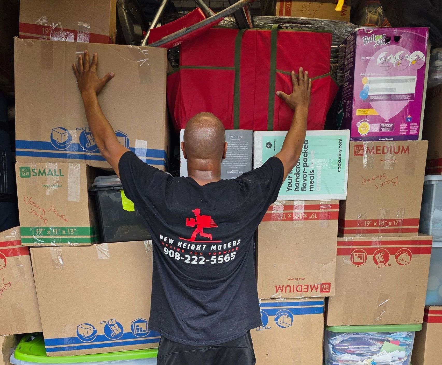 Man with arms raised in front of stacked boxes, truck interior. He's wearing a black shirt and facing away.