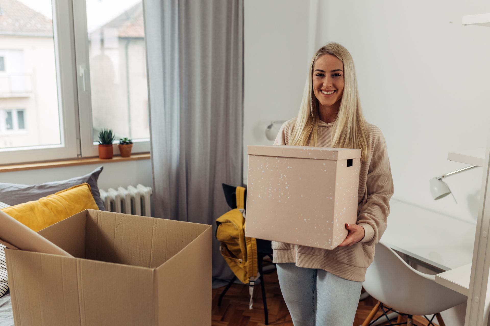 Blonde woman smiling, holding a box indoors, possibly moving. Boxes and a window are visible.
