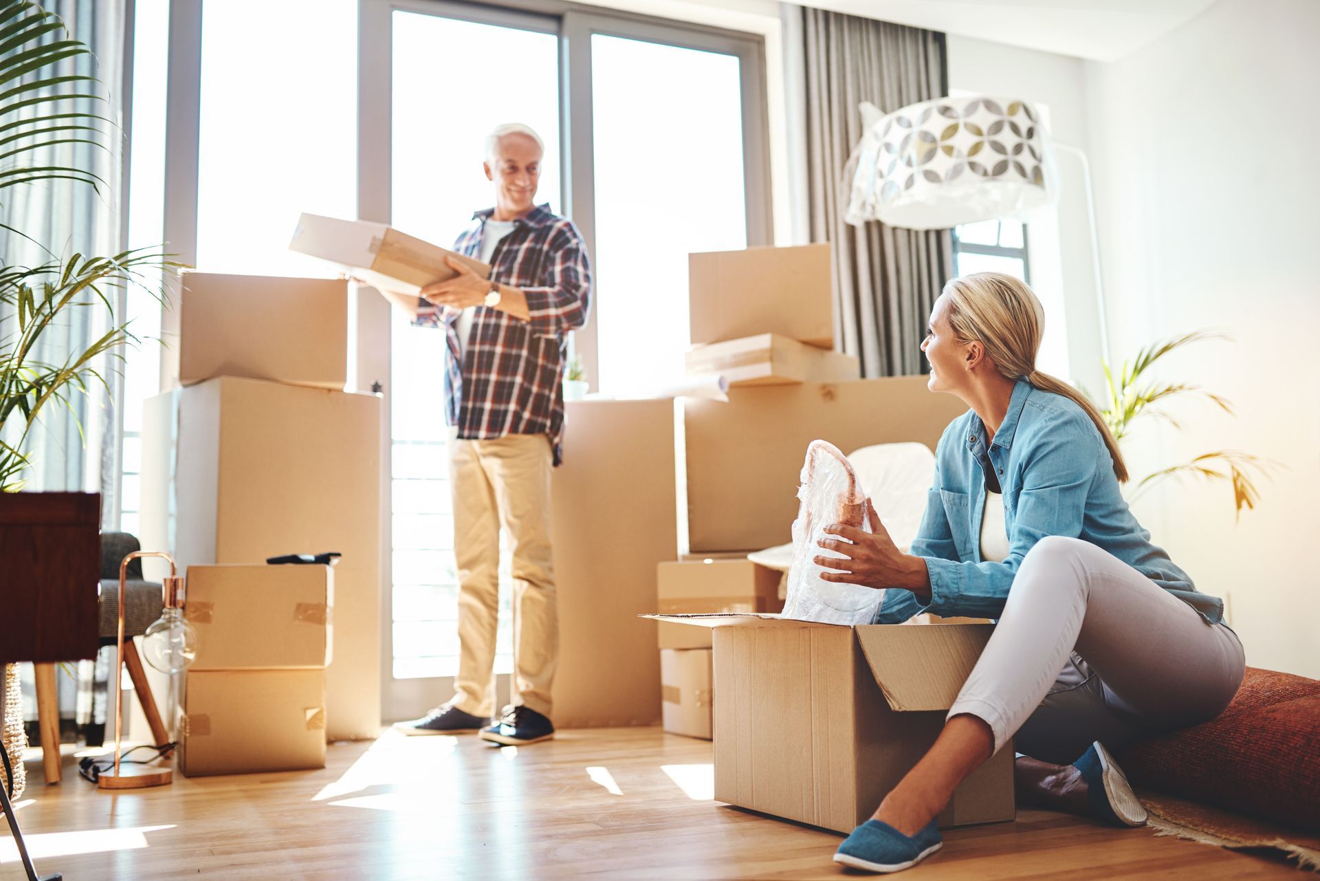 Couple unpacking boxes in a new home, man holding box, woman seated, surrounded by cardboard boxes.