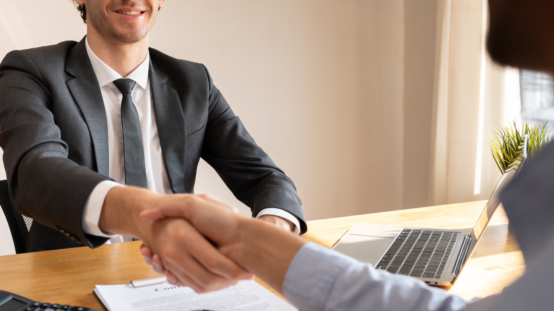 Man in a suit smiles, shaking hands with a person at a desk with a laptop and documents.