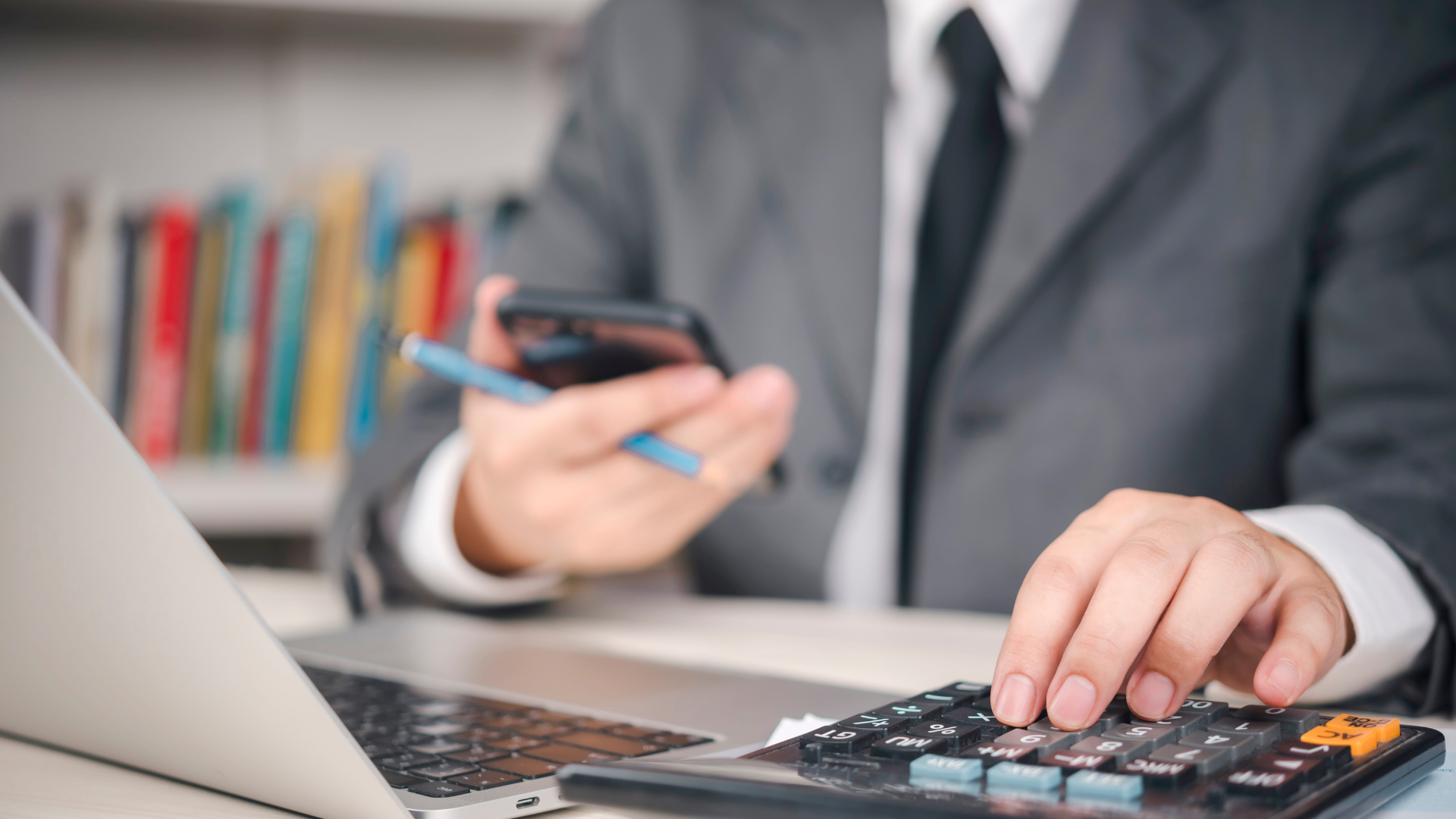 Person in suit using a calculator, holding a phone, and working on a laptop in an office setting.