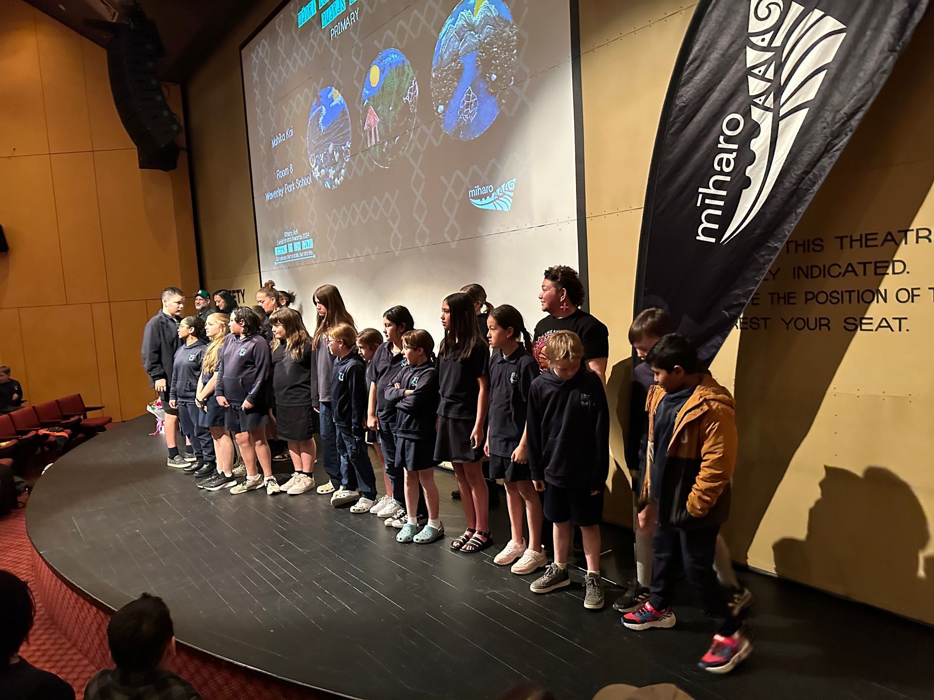 A group of children are standing on a stage in front of a large screen.