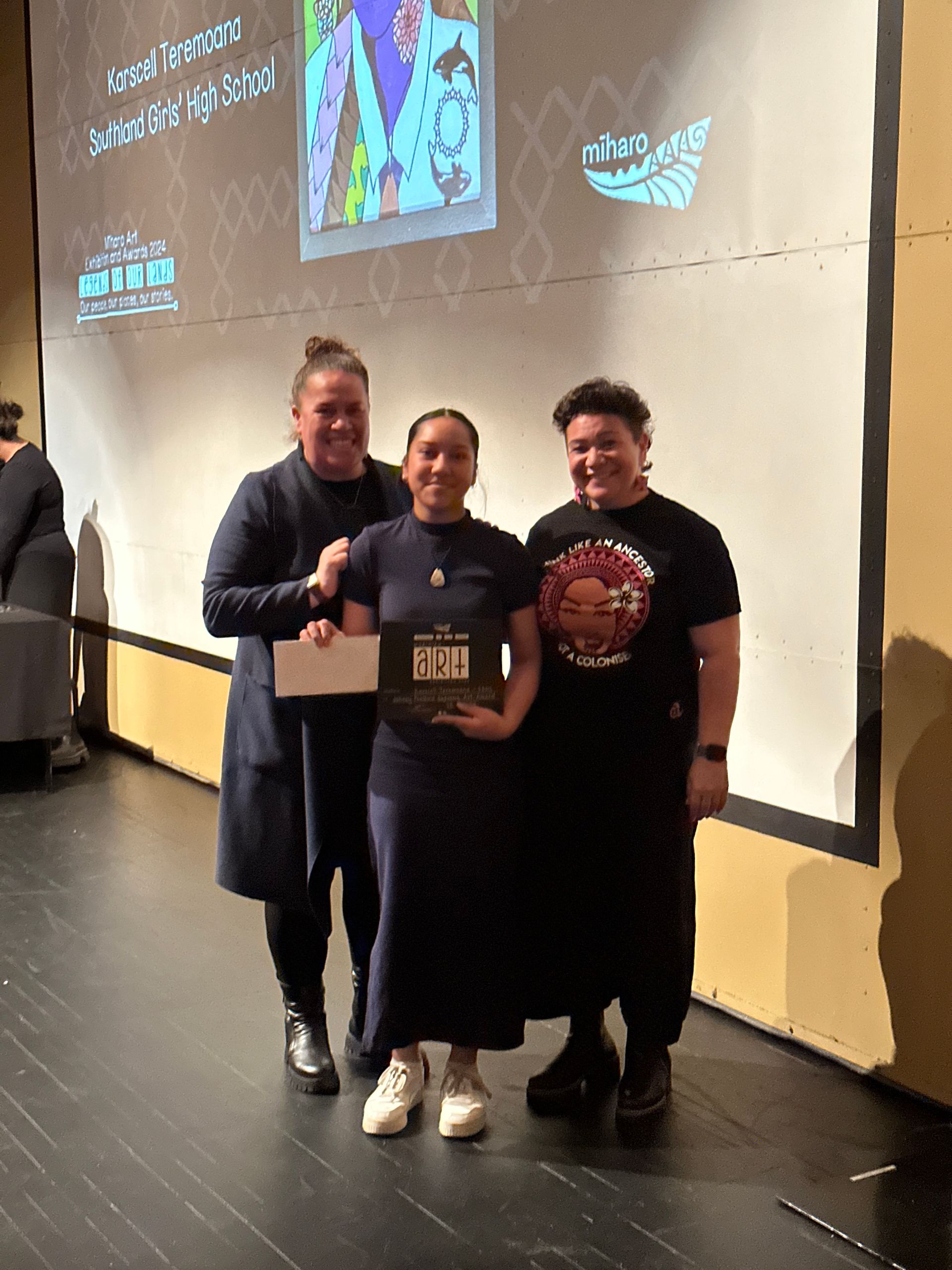 Three women are posing for a picture in front of a large screen.