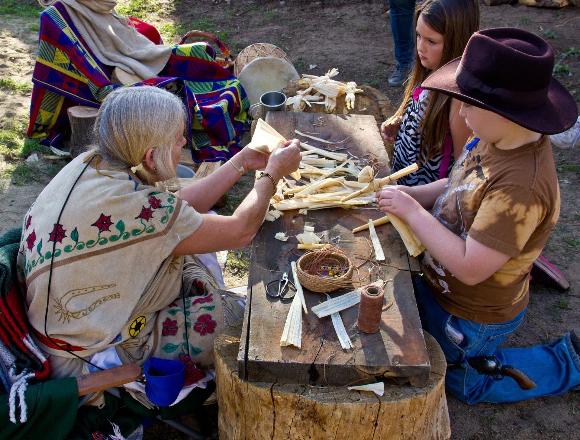 People make corn husk dolls around a wood-stump table