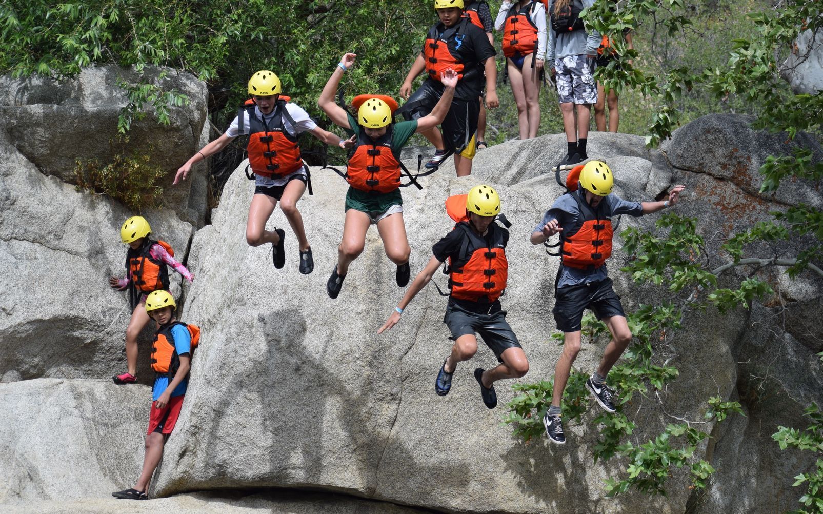 People in helmets and life vests jump off a rock into water. Sunny outdoor setting.