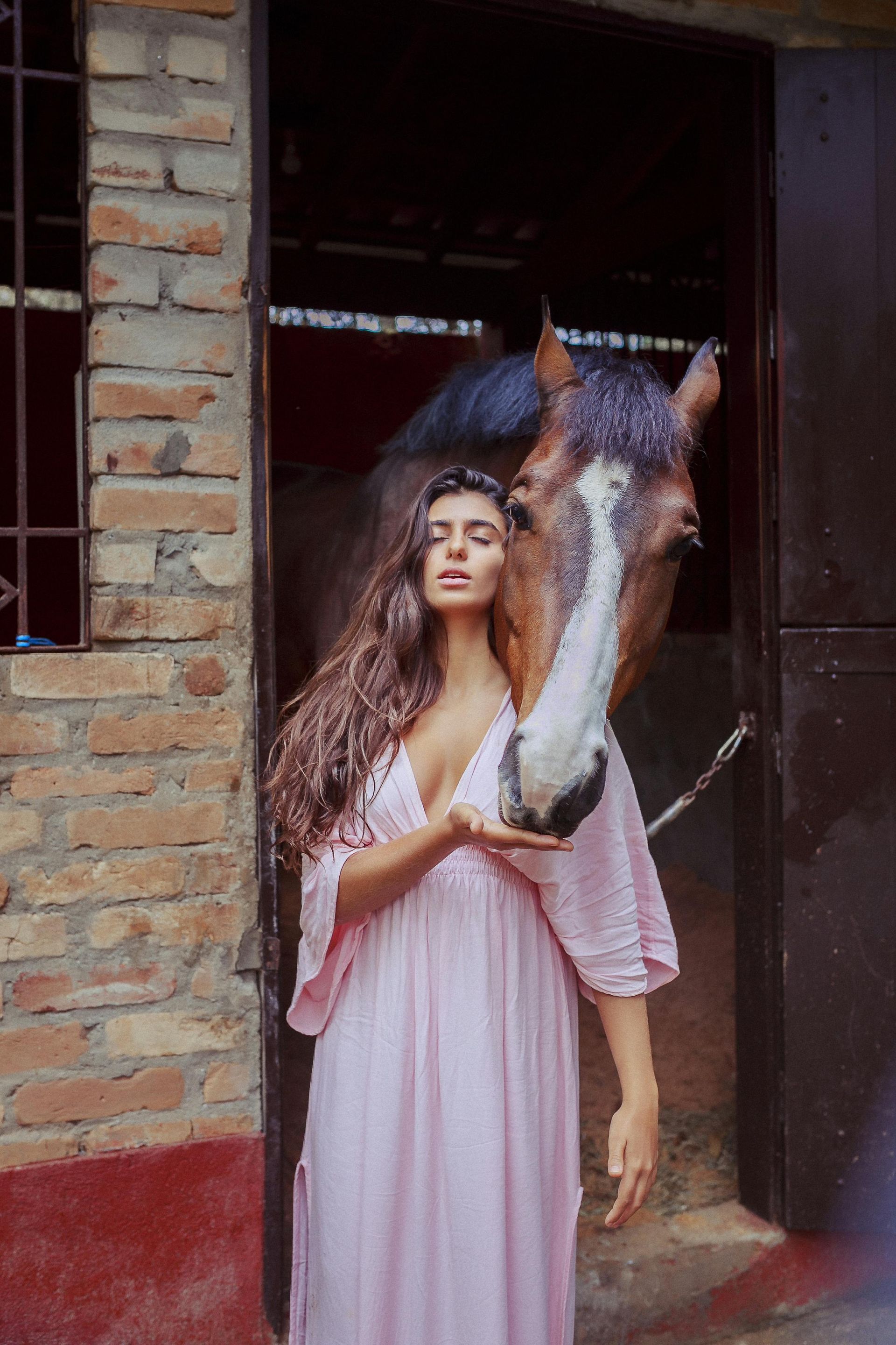 A woman stood by a stable with a horse who has their head over her shoulder. 