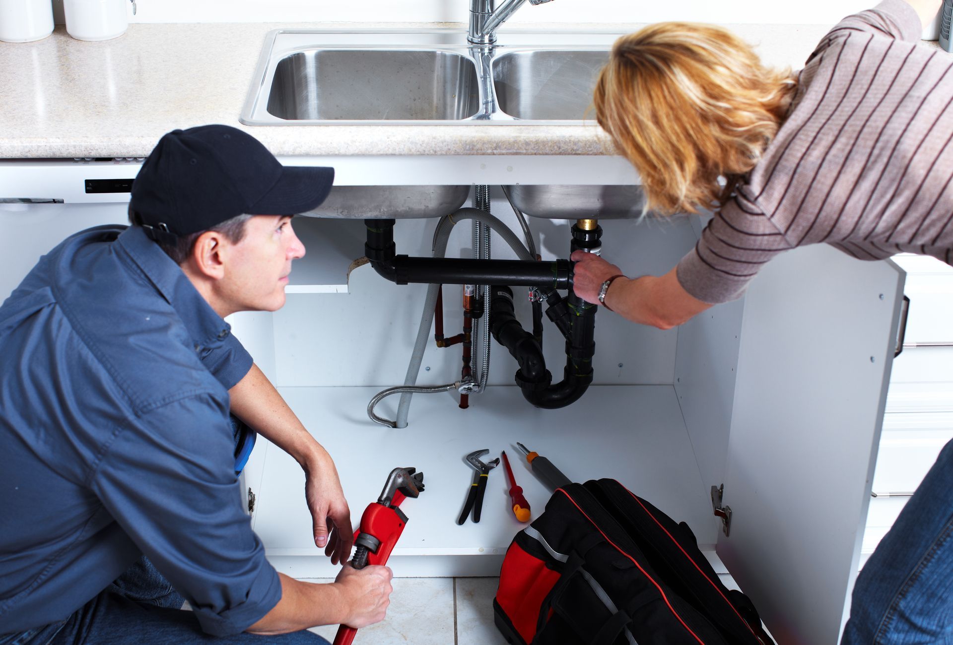 A professional plumber fixes sink pipes while the client points out the issue A professional plumber fixes sink pipes while the client points out the issue