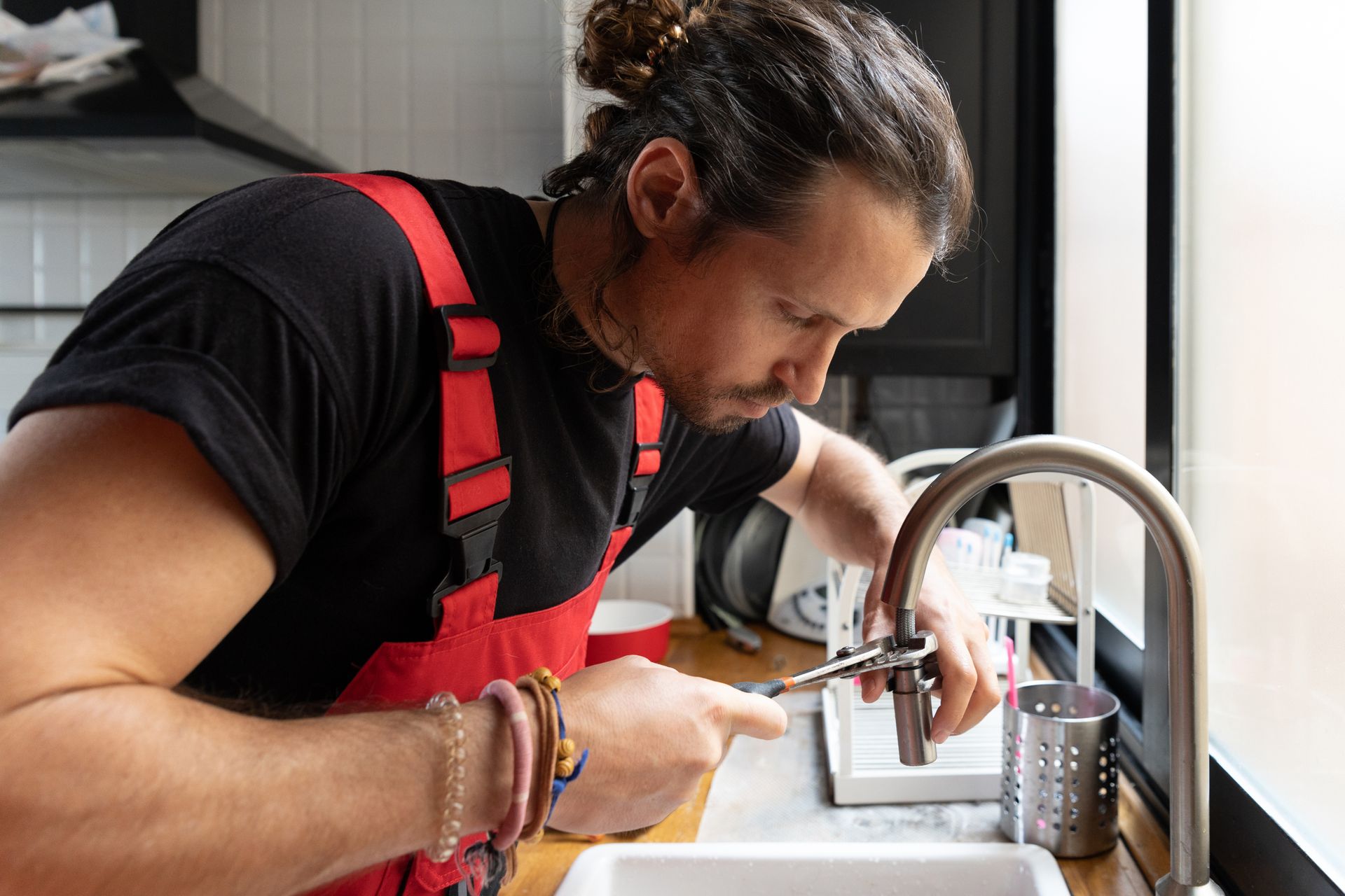A male plumber in red overalls is fixing a kitchen faucet with a wrench.