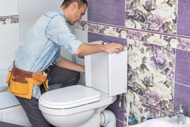 Person repairing toilet tank in a pink floral-tiled bathroom.