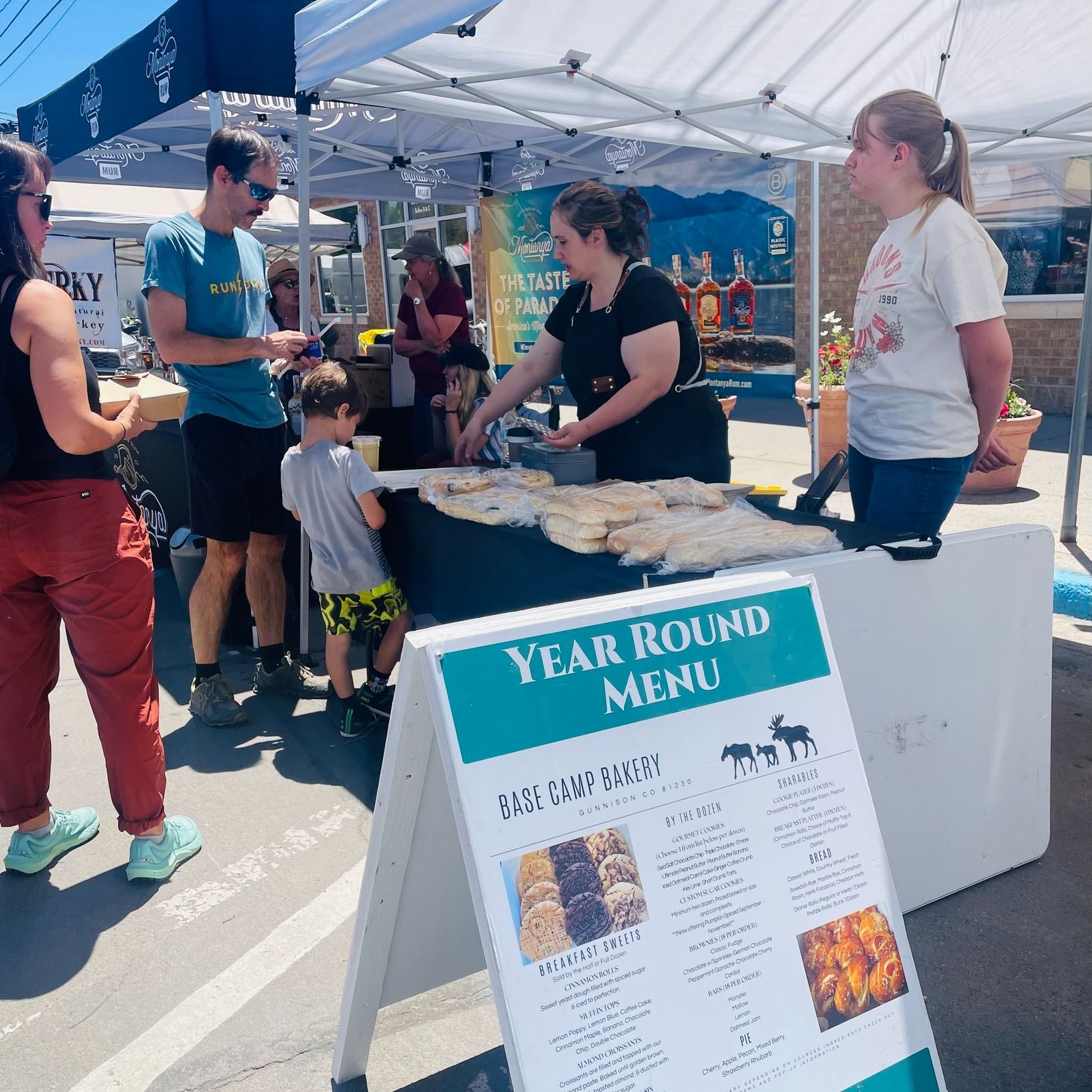 People at a food market stand with a menu; baked goods displayed. Base Camp Bakery Gunnison, Colorado https://share.google/AgqUzUKNzsb0ThlP5, https://www.facebook.com/BaseCampBakery1, https://www.instagram.com/basecampbakery_81230