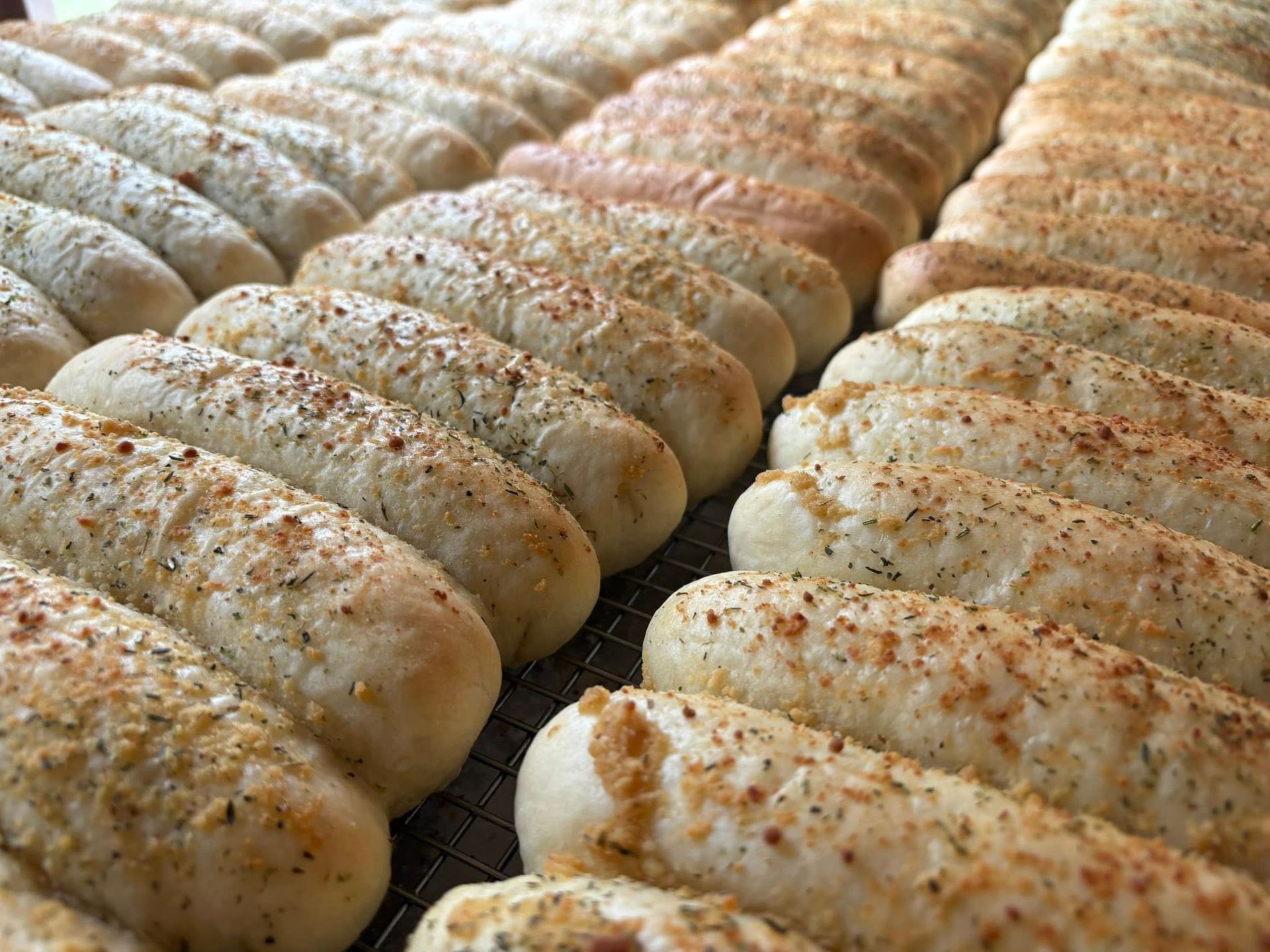 Rows of baked breadsticks, some sprinkled with seasonings, on a cooling rack.