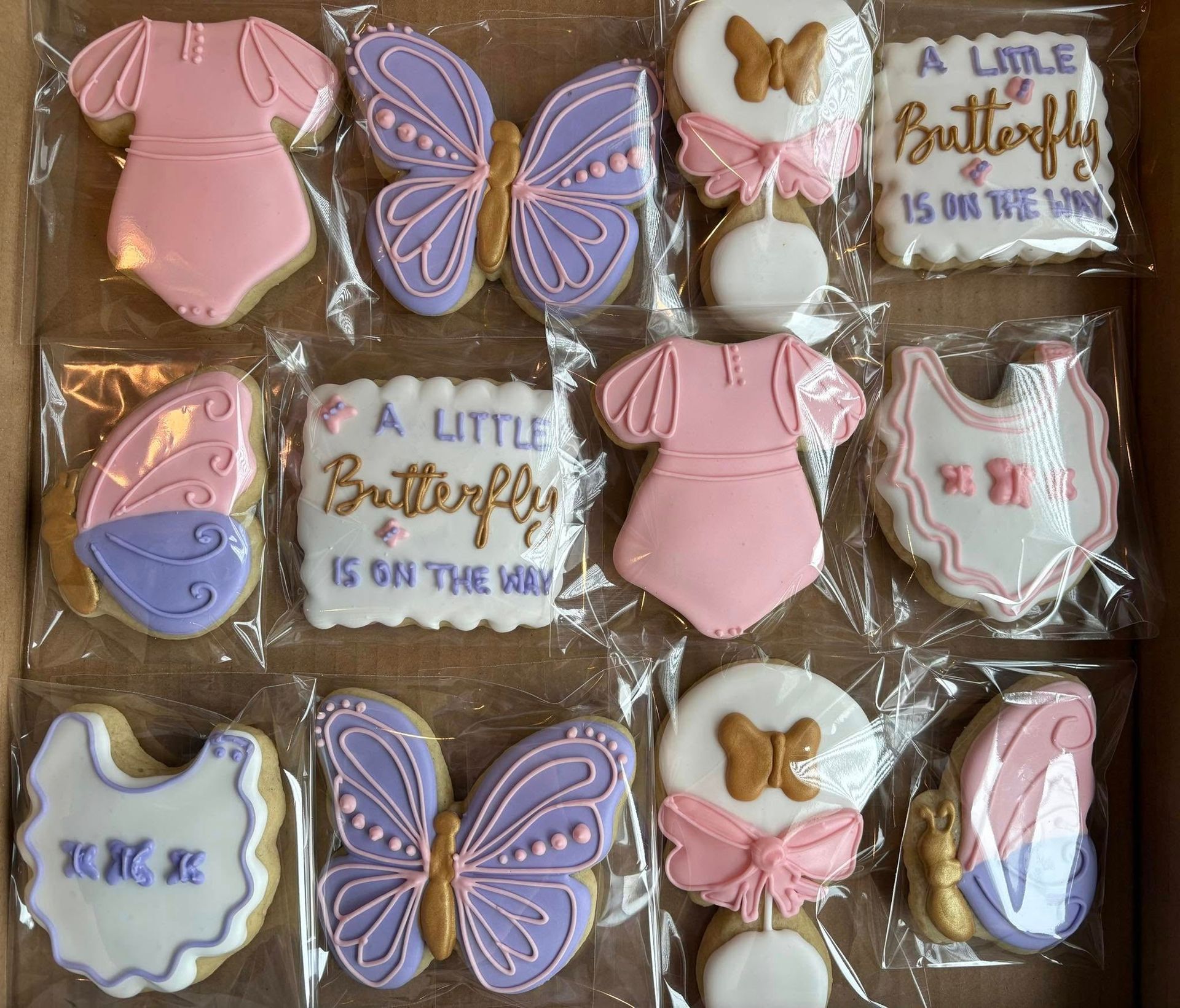 Box of decorated baby shower cookies: butterflies, onesies, bibs, and rattles in pink, purple, and gold.