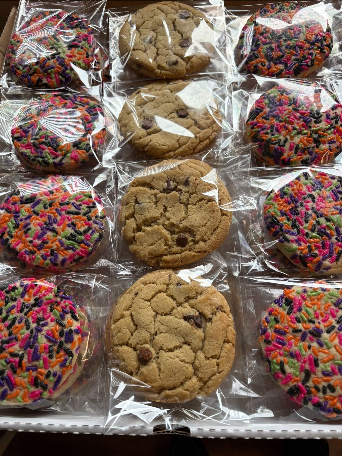 Cookies in clear plastic wrappers arranged in a box. Some are sprinkled, others have chocolate chips.