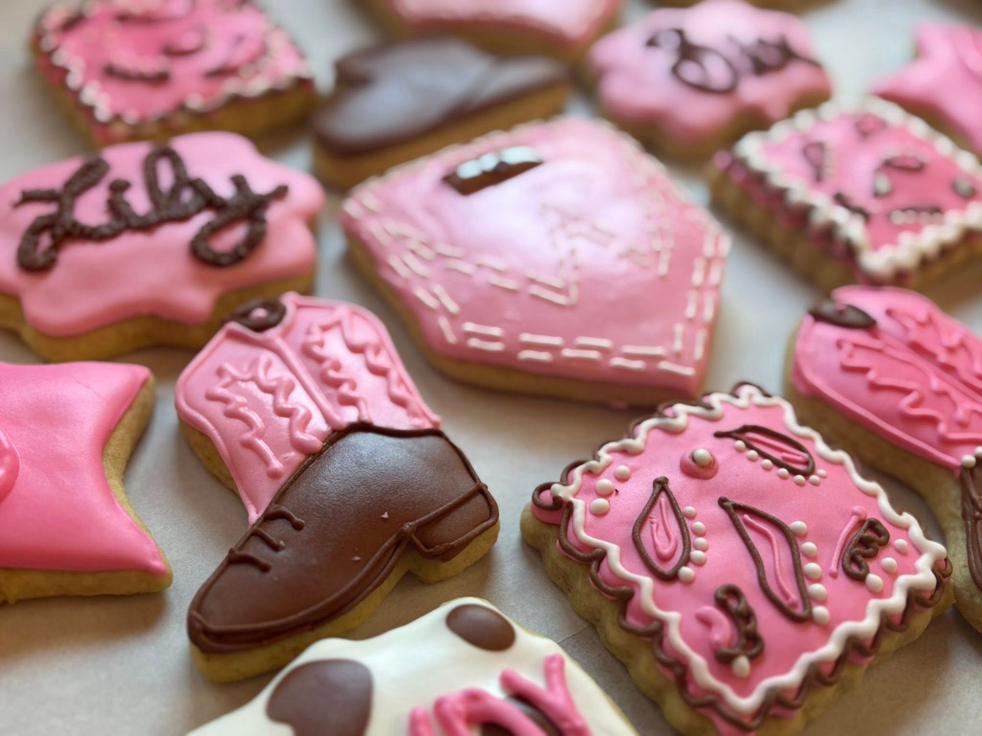 Pink frosted decorated cookies, including cowboy boot, with names, and floral and geometric designs.
