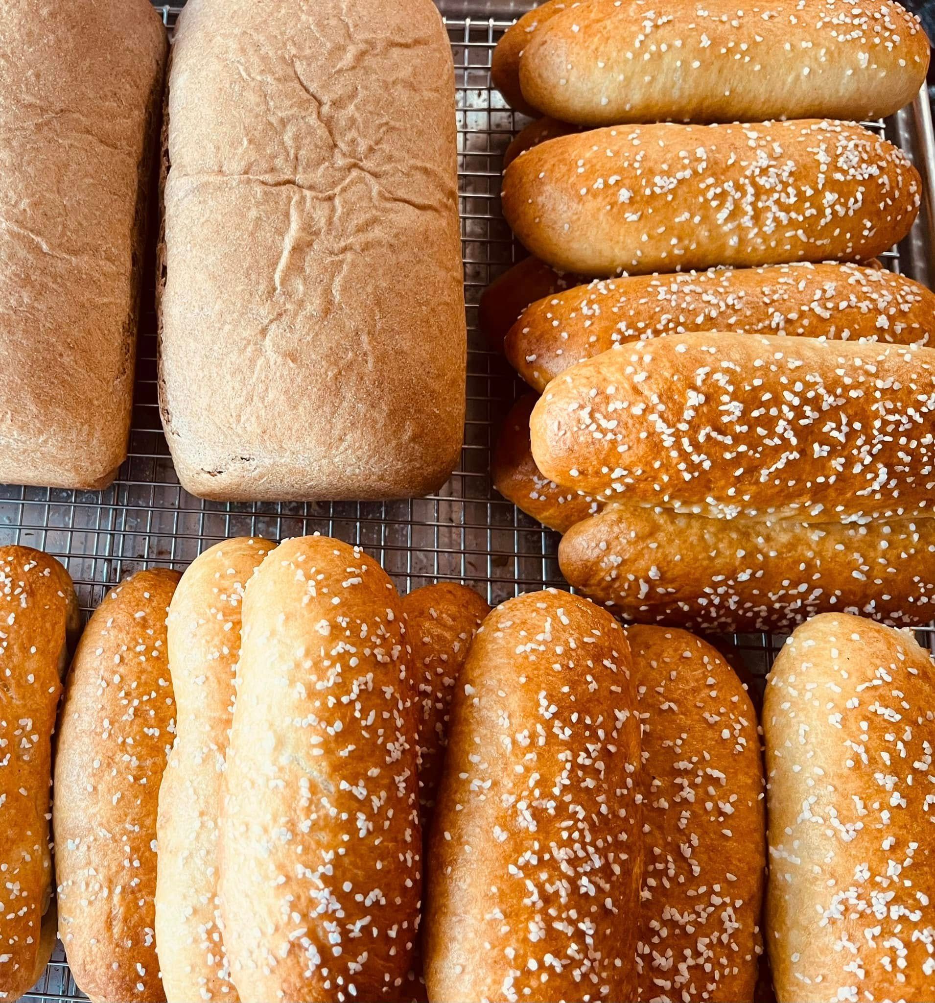 Freshly baked bread rolls and loaves on a wire rack, some with sesame seeds.