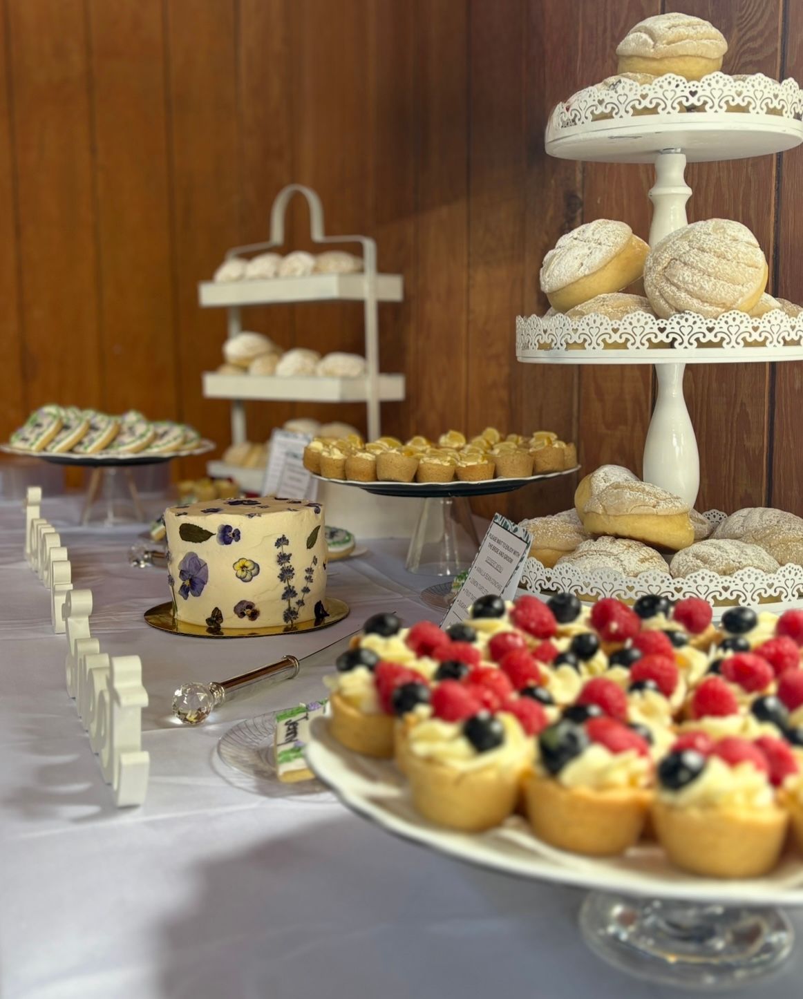 Dessert table: tiered stands with cookies, tarts with berries, cake, and mini pastries on a white tablecloth.