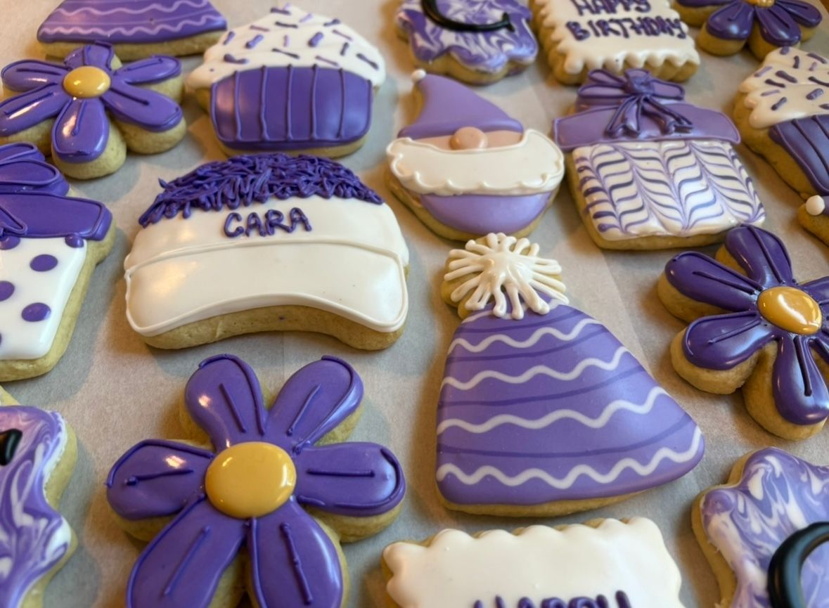 Purple frosted cookies: cupcakes, flowers, hats, a gnome, and a gift box, decorated for a celebration.