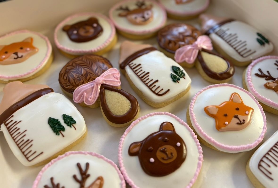 Box of decorated baby-themed cookies: bottles, rattles, fox, bear, and deer faces, white and pink frosting.