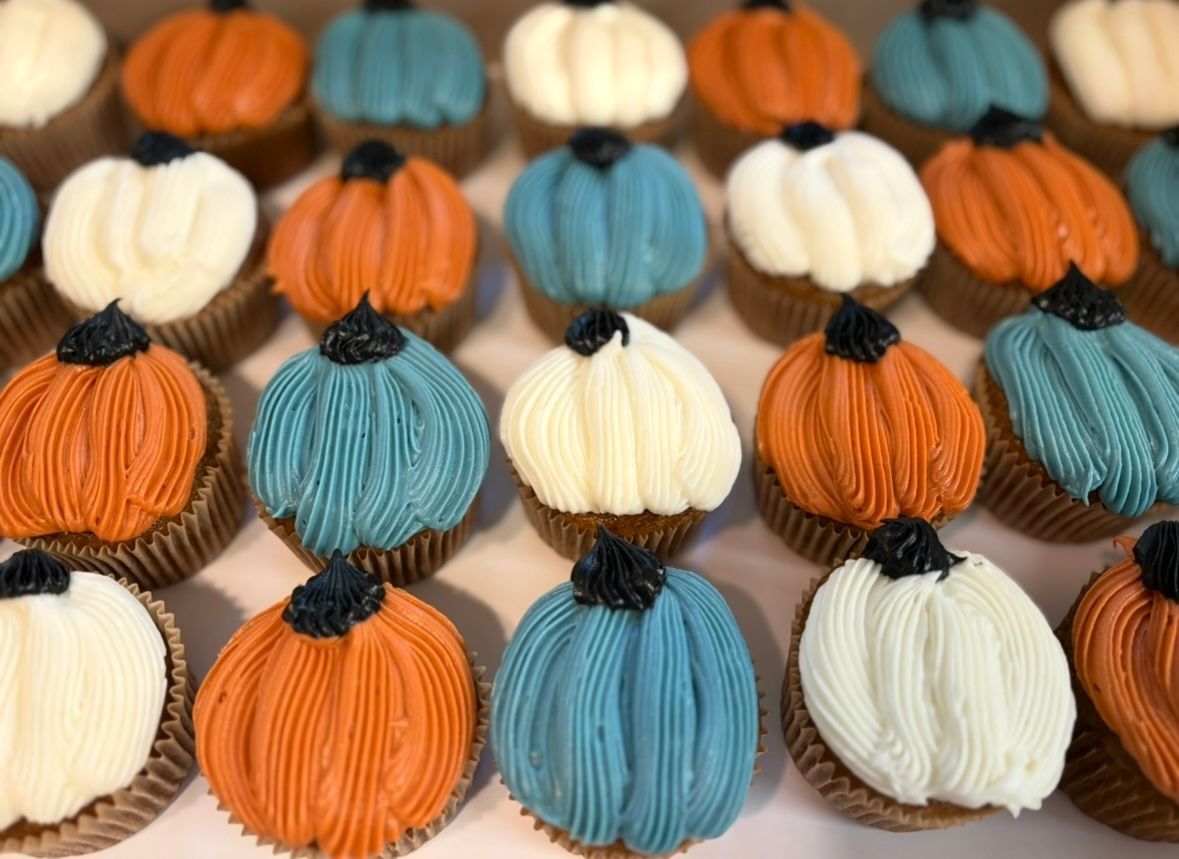 Cupcakes decorated as pumpkins with orange, white, and teal frosting, in a box.