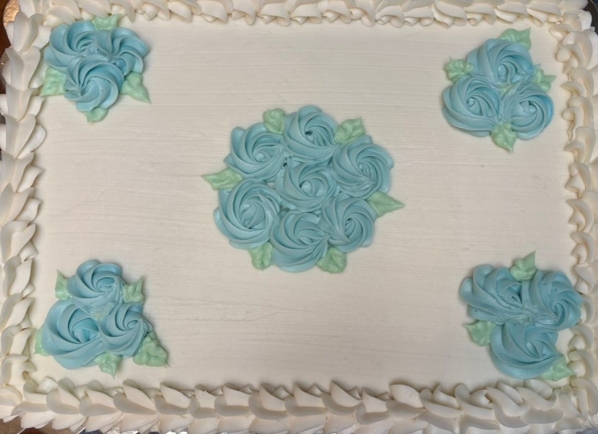 Rectangular cake with white frosting and blue frosting roses; white scalloped border.