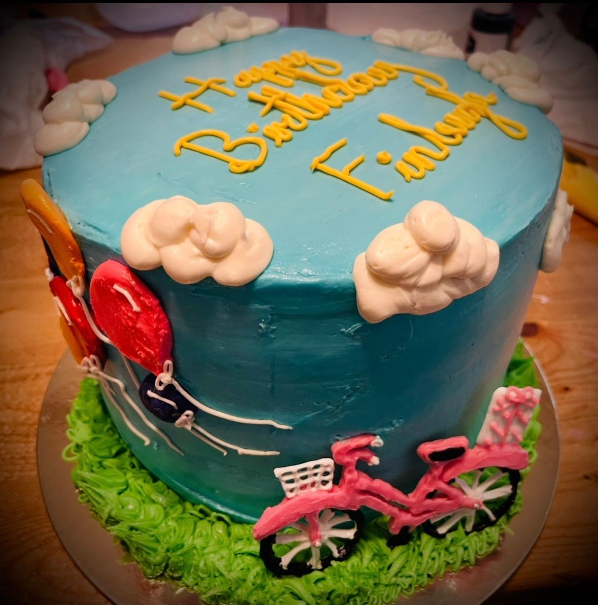Birthday cake with blue frosting, clouds, balloons, and pink bicycle. 