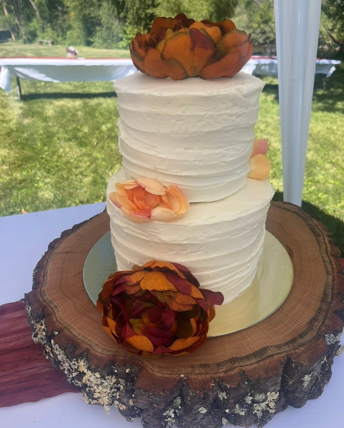 Two-tiered white frosted cake with fall-colored flowers on a wood slice. Outdoors, sunny setting.