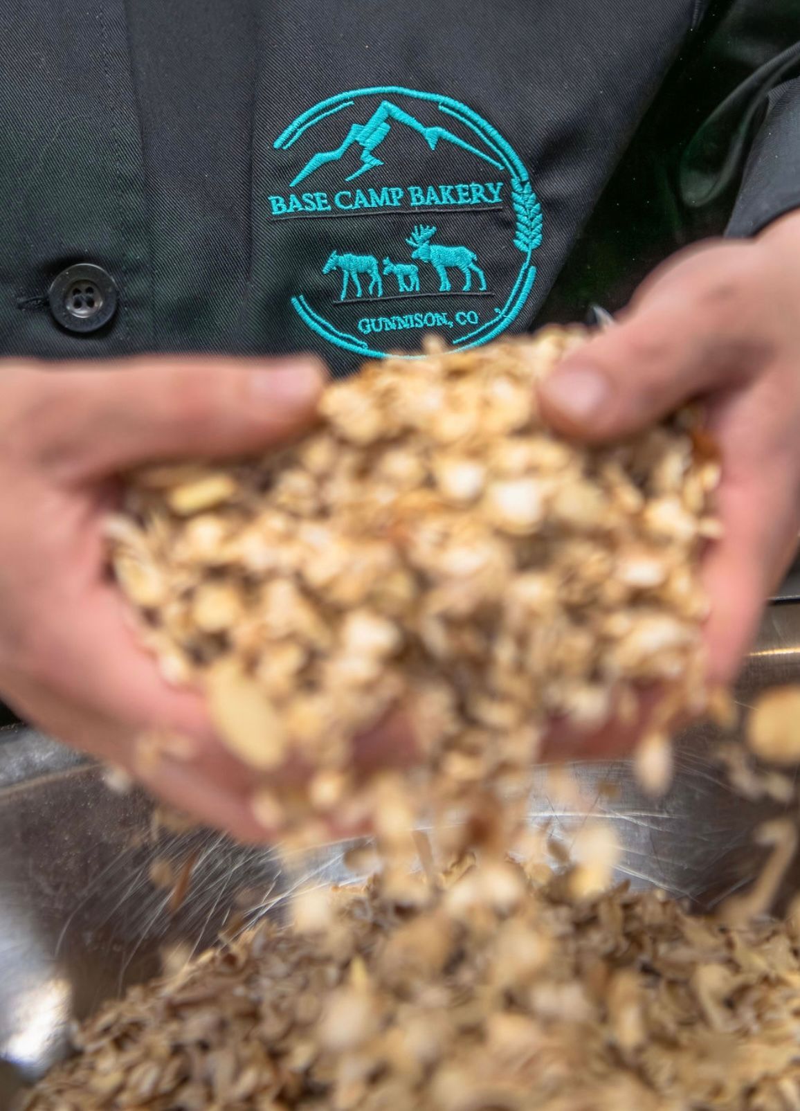 Person holding chopped nuts over a bowl, wearing a black jacket with a Base Camp Bakery logo.