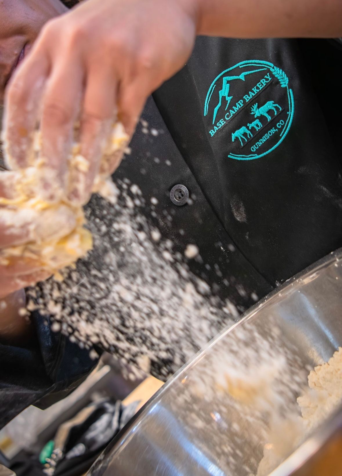 Person in black apron kneads dough, flour flying; logo reads "Drake Camp Bakery, Farmington, CO."