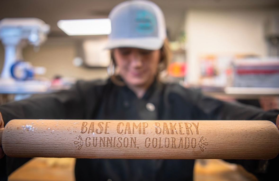 Baker holding rolling pin, "Base Camp Bakery, Gunnison, Colorado" is engraved on it.
