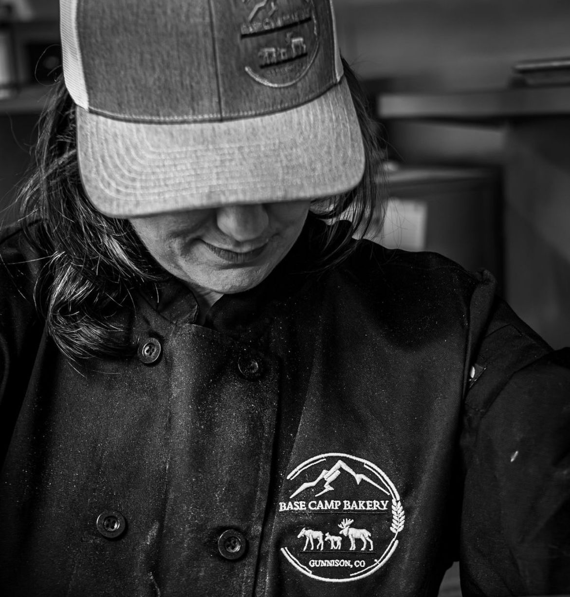 Baker wearing hat and jacket with bakery logo, focused on their work in a kitchen.