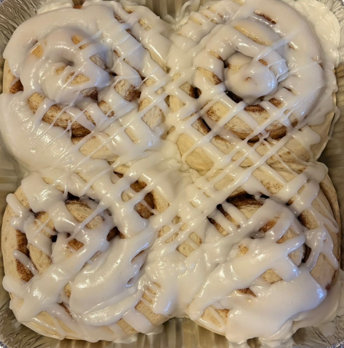 Four frosted cinnamon rolls in a foil pan, drizzled with white icing.