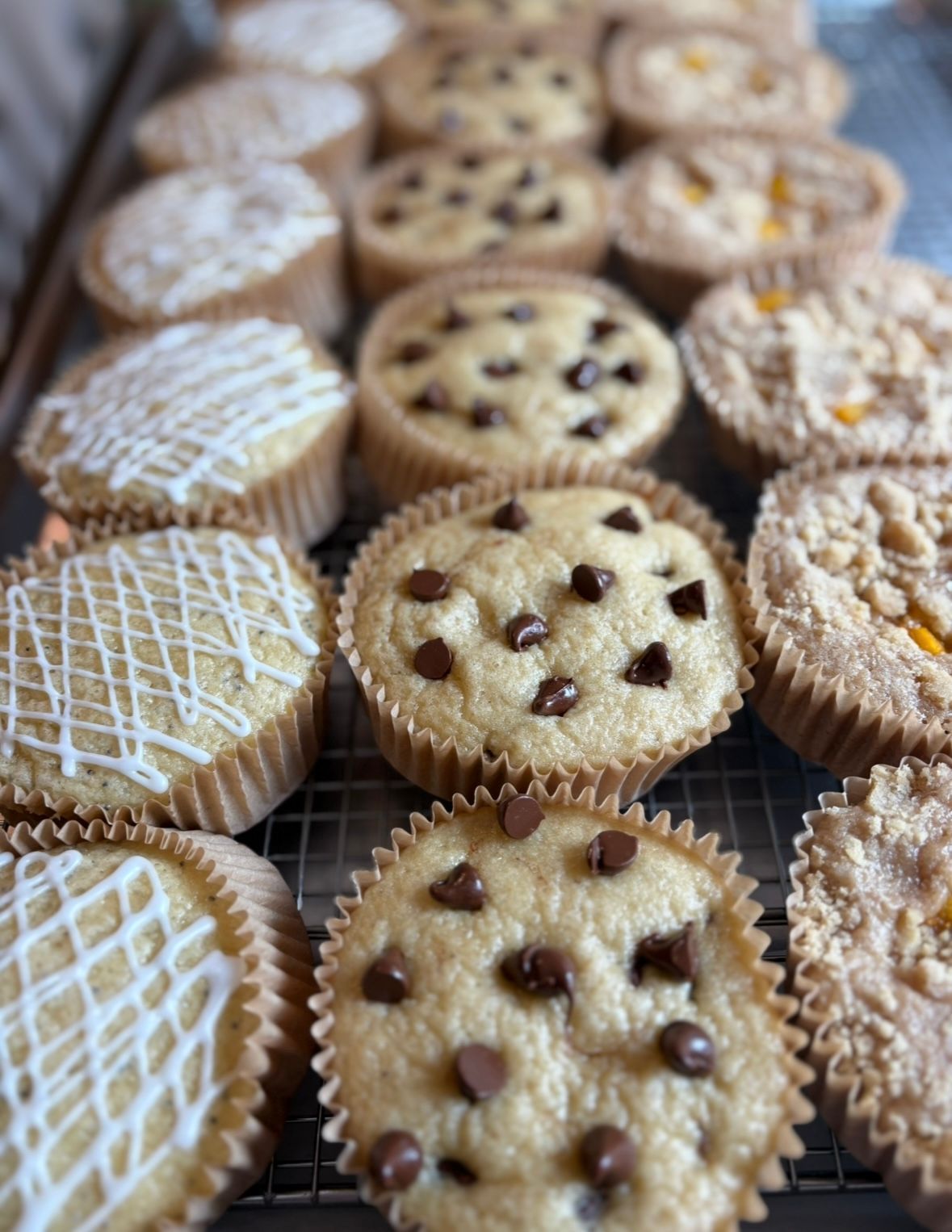 Rows of muffins, some with chocolate chips, icing, and crumble toppings, on a wire rack.
