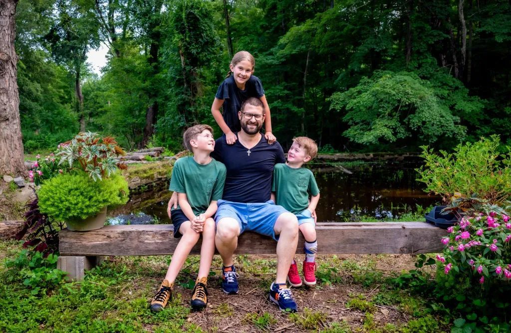 A man sits on a bench with three children. The eldest is on his shoulders. Green foliage surrounds them.