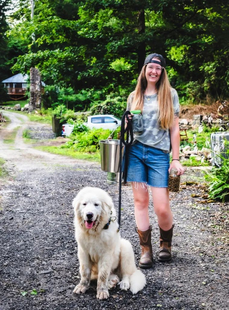 Woman with long hair, wearing boots and hat, stands with a large white dog on a gravel path, holding a leash.