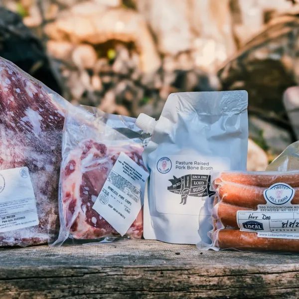 Various cuts of raw meat packaged outdoors on a wooden surface; includes sausages, and pork bone broth.