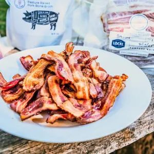 Plate of crispy bacon on a wooden surface, with bags of pork products in the background.