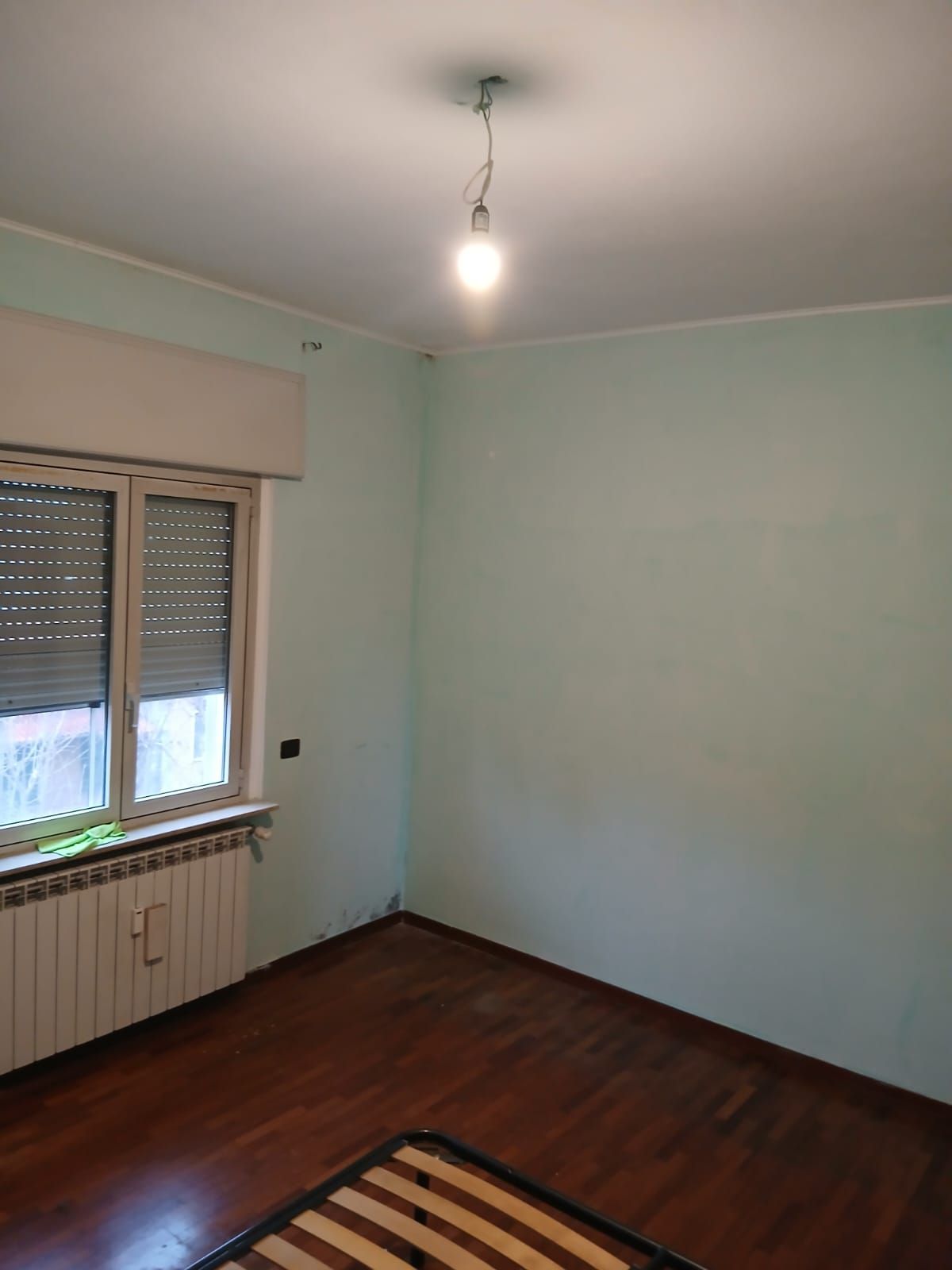 Camera da letto vuota con pavimento in legno marrone, pareti verde chiaro, finestra, termosifone e lampadina appesa.