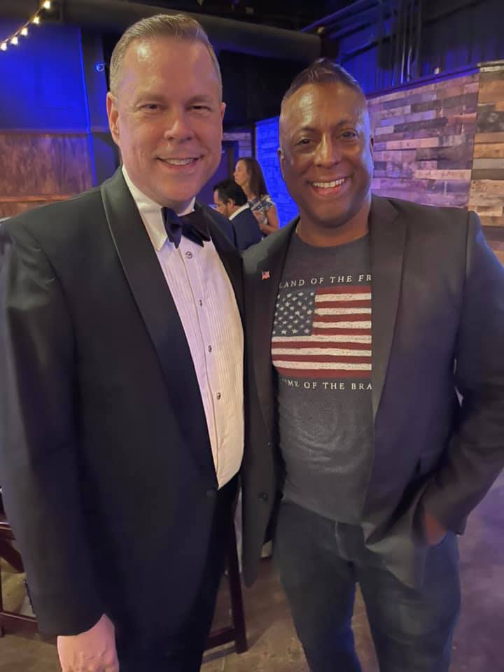 A man in a tuxedo and a man in an american flag shirt are posing for a picture.