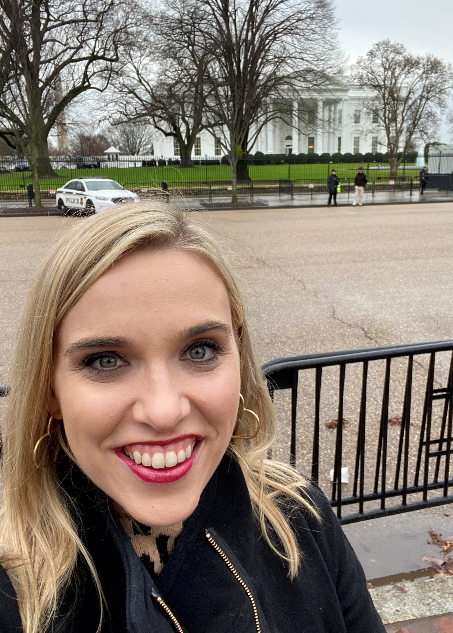 A woman in a black coat is smiling in front of the white house.