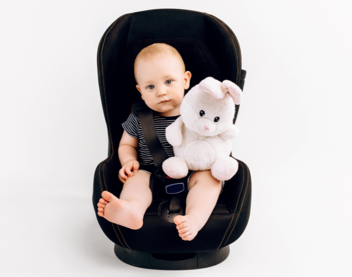 Bambino nel seggiolino auto nero con coniglietto di peluche bianco, su sfondo bianco.