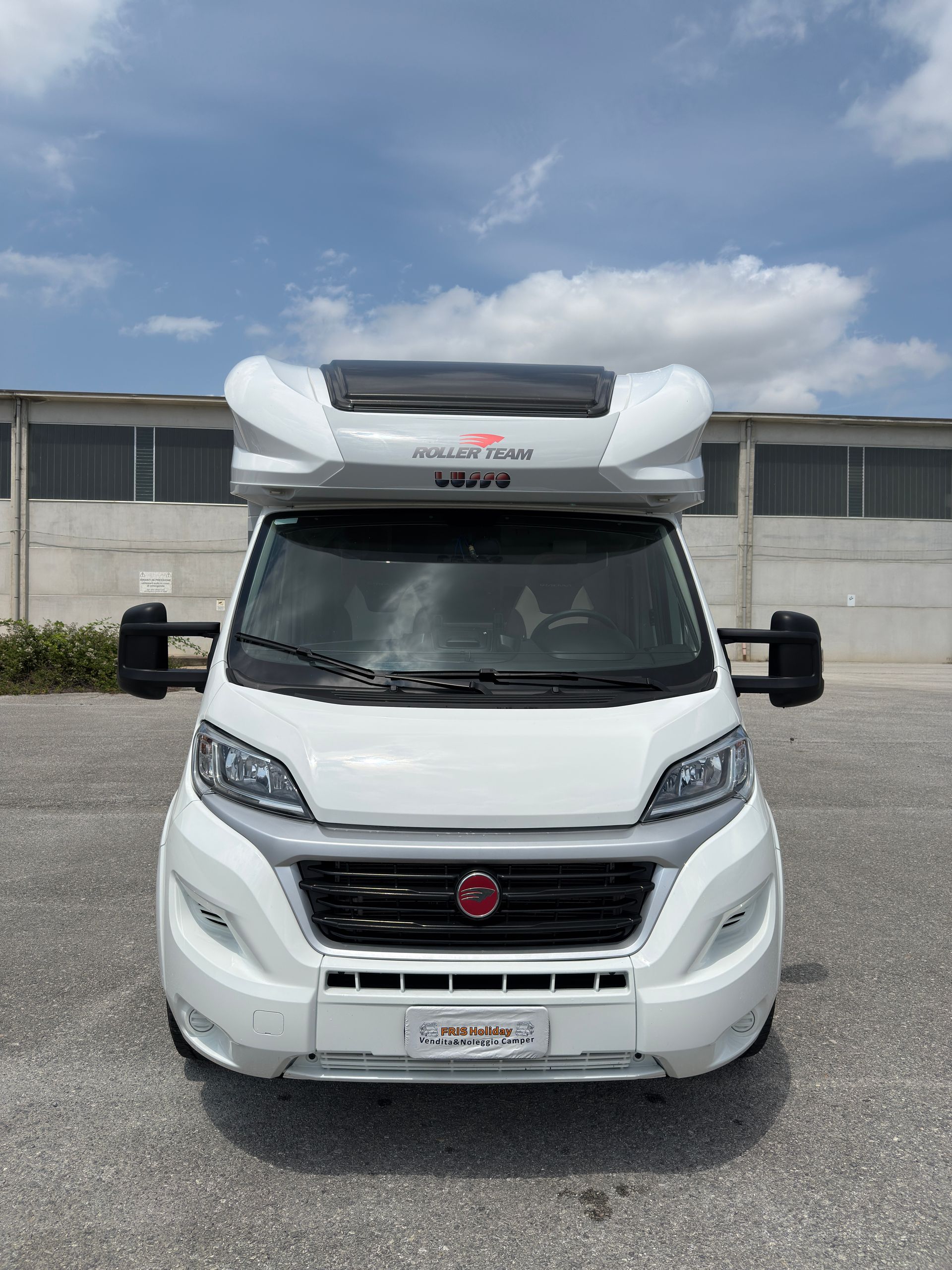 Camper bianco con tetto arrotondato, parcheggiato all'aperto contro un cielo e un edificio.