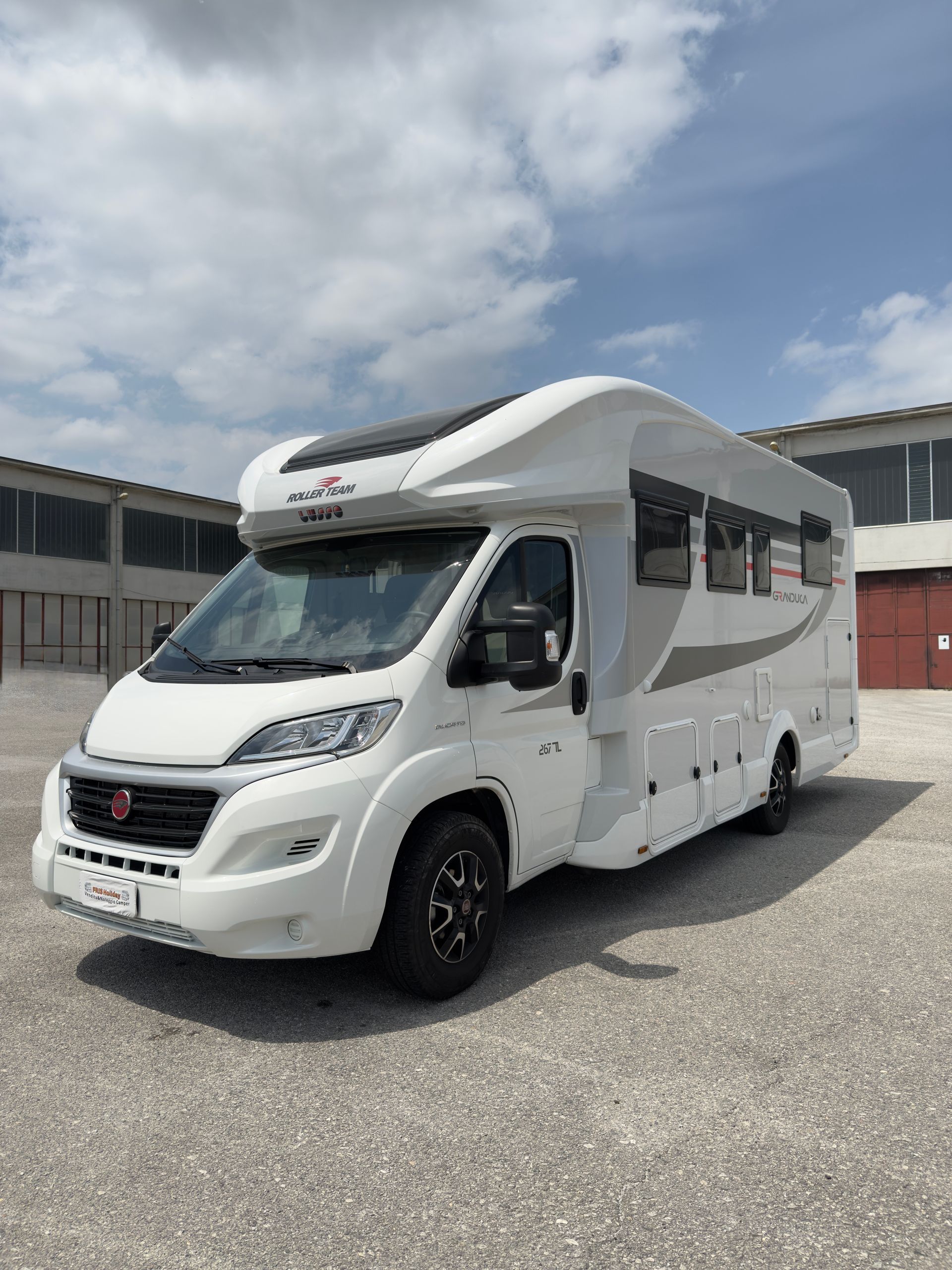 Camper bianco e grigio parcheggiato all'aperto davanti a un edificio in mattoni in una giornata di sole.