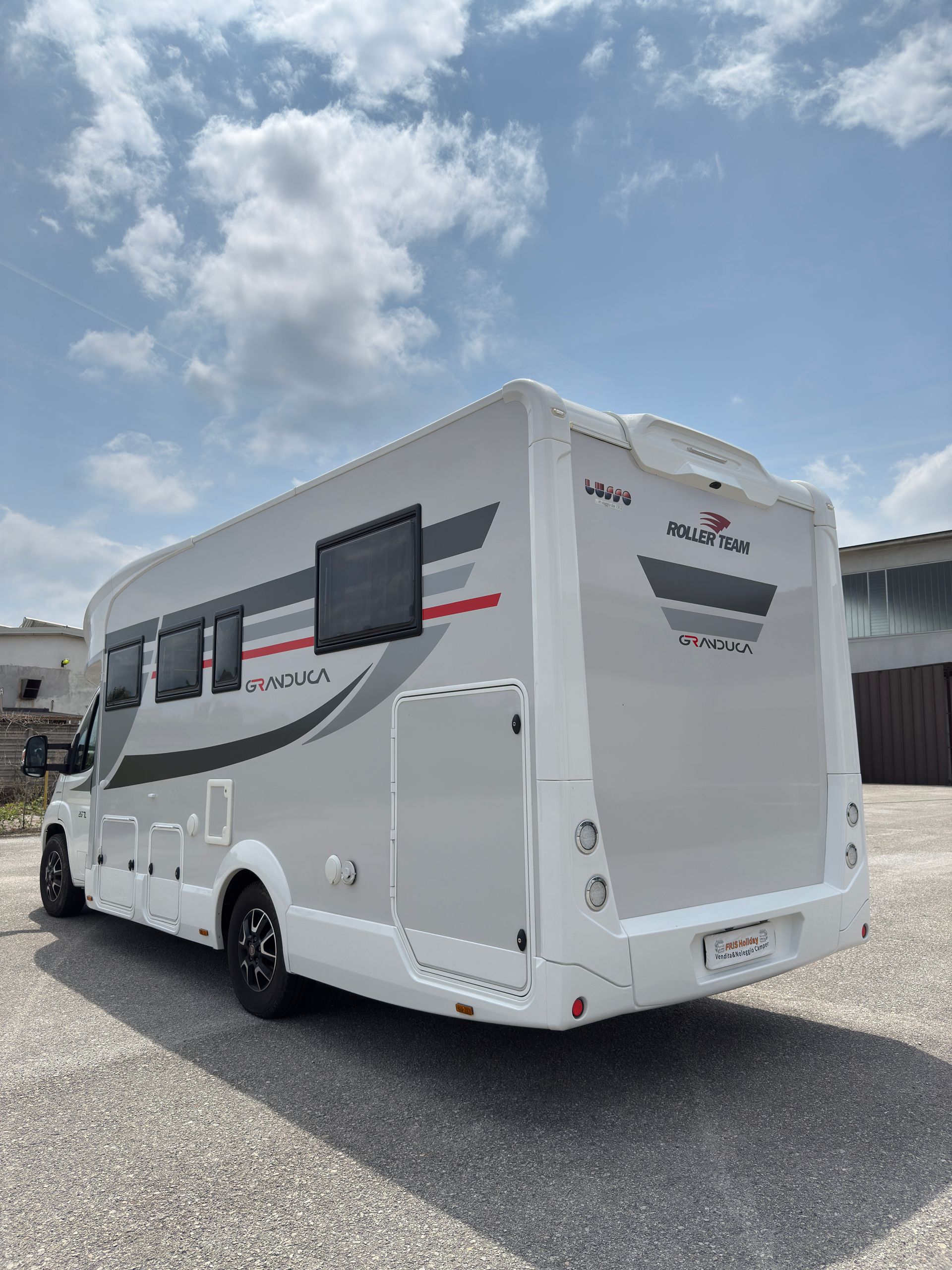 Camper bianco e grigio parcheggiato all'aperto su un terreno ghiaioso sotto un cielo nuvoloso.