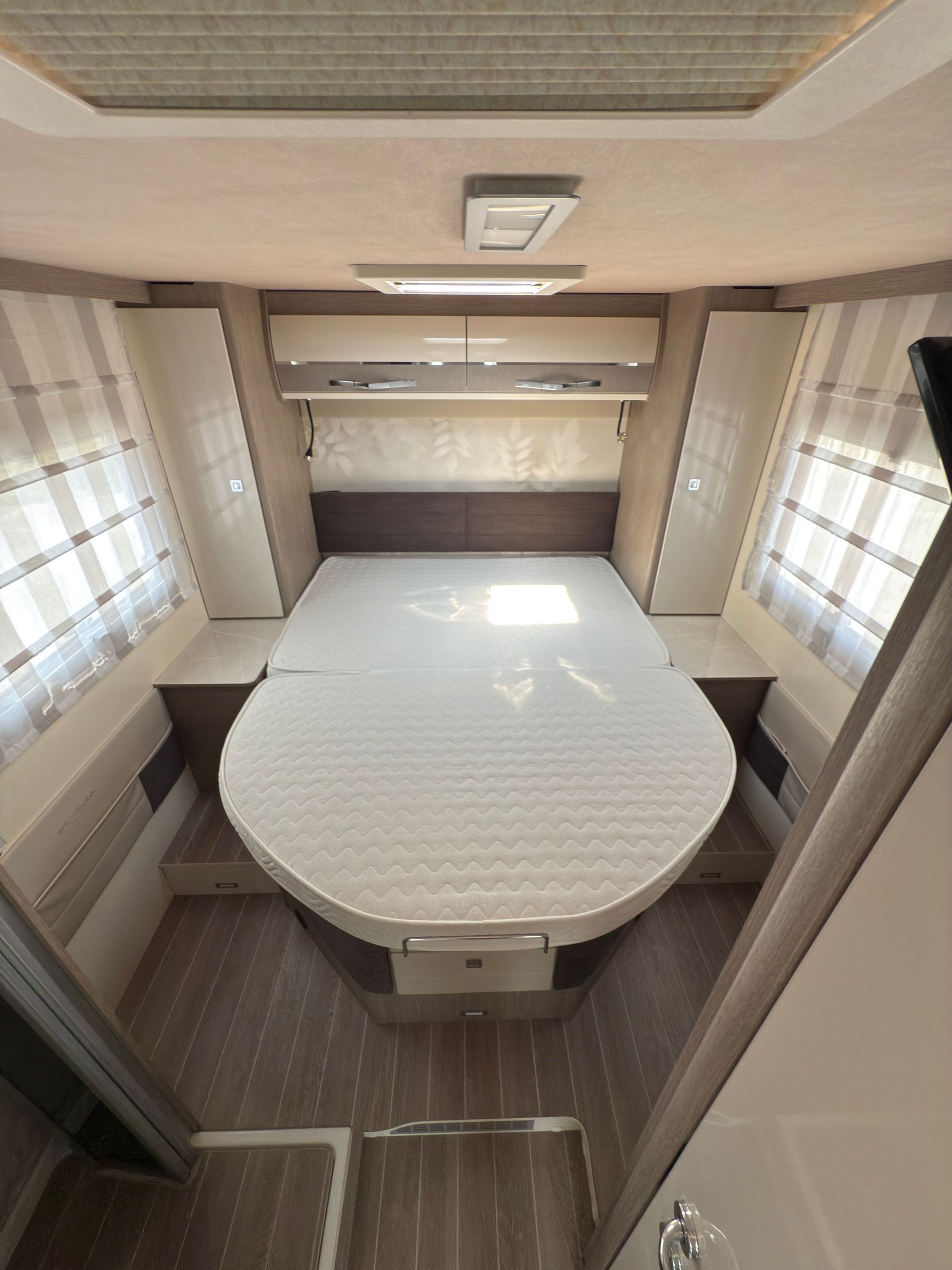 Vista interna di una camera da letto di un camper con un letto grande, armadi e una finestra con persiane.