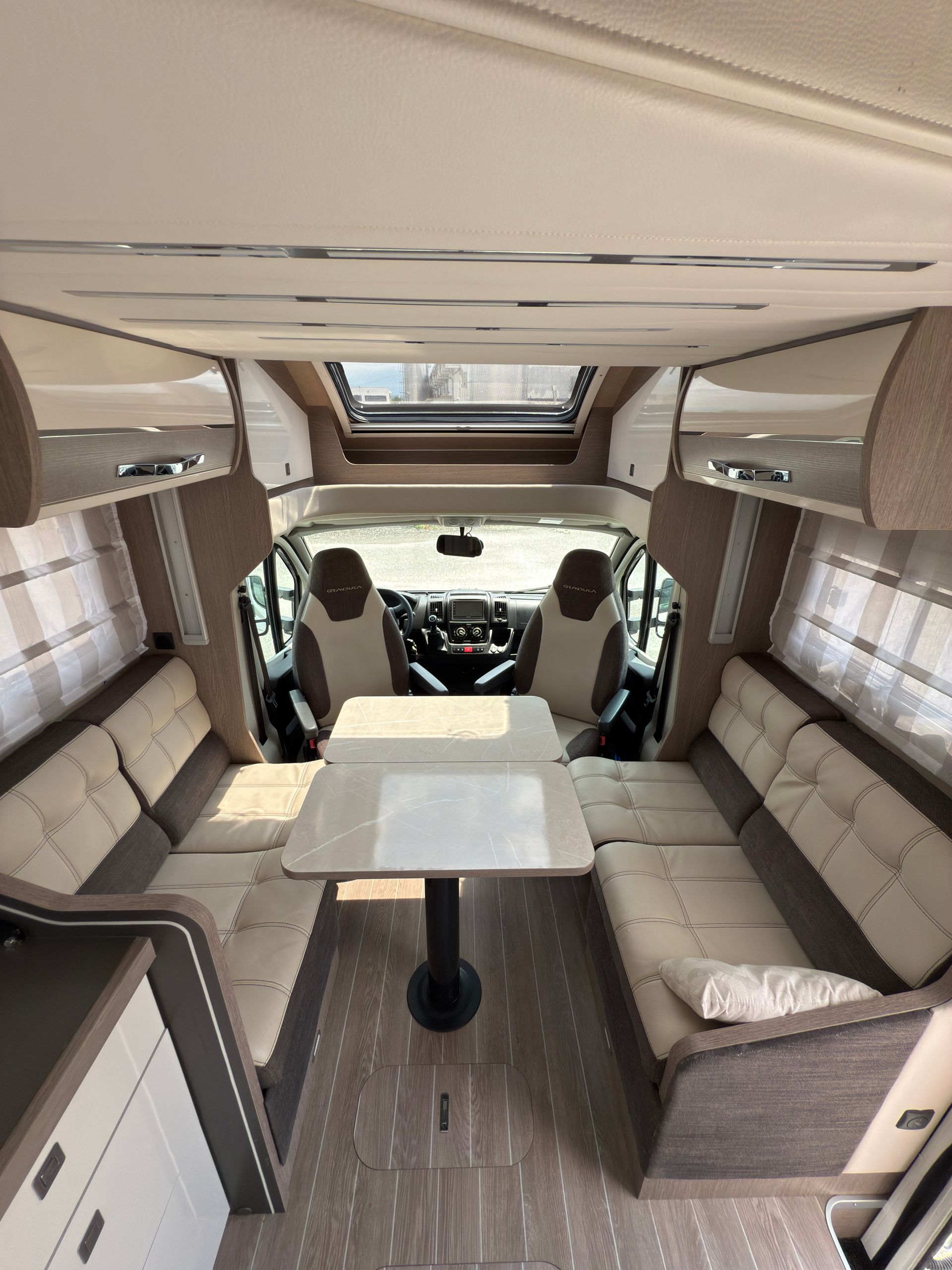 Interno di un camper, con zona di seduta, tavolo e posto guida visibili. Colori chiari, layout aperto.