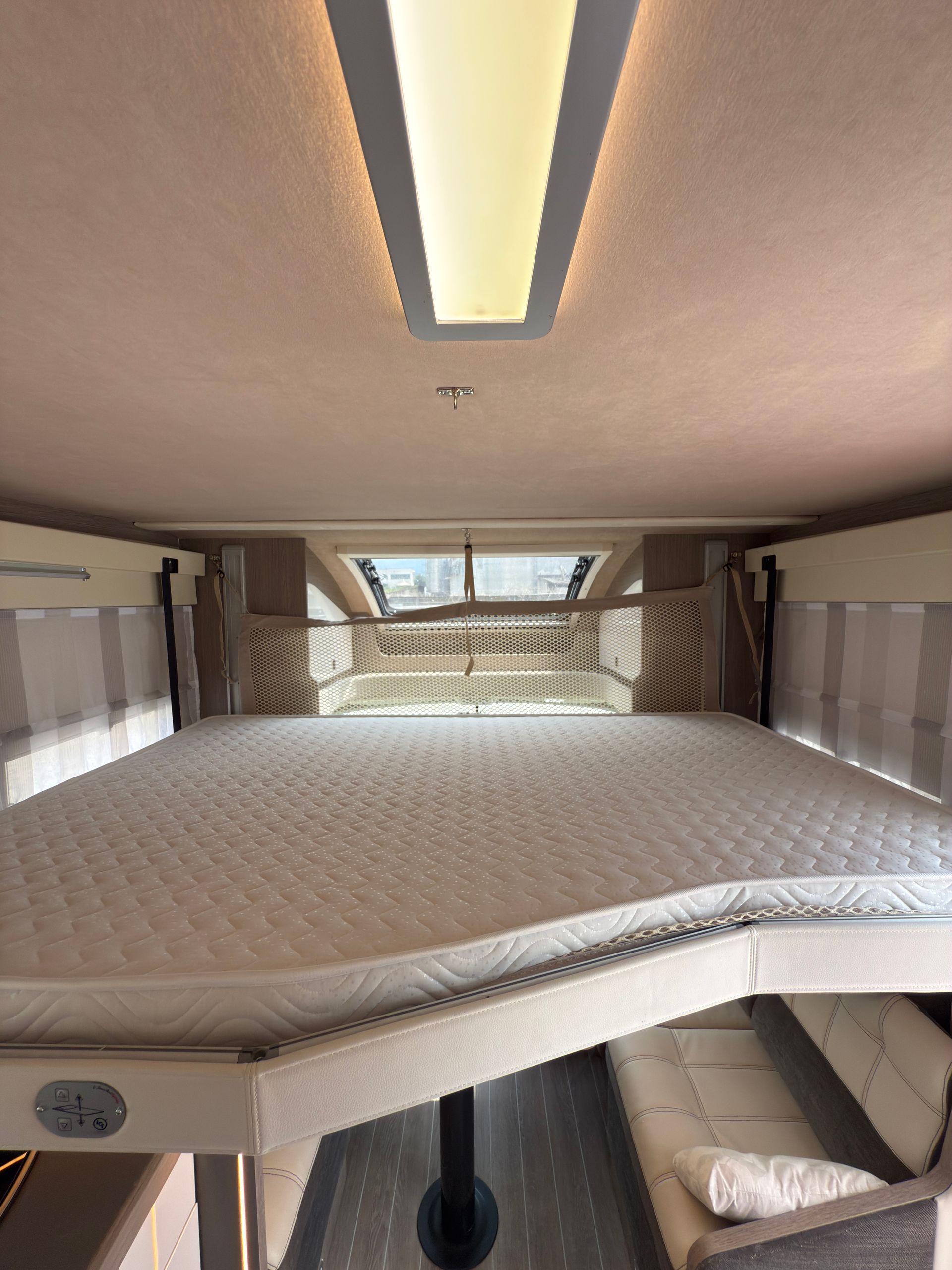 Interno del camper: letto sopra la zona salotto, combinazione di colori beige e bianco.