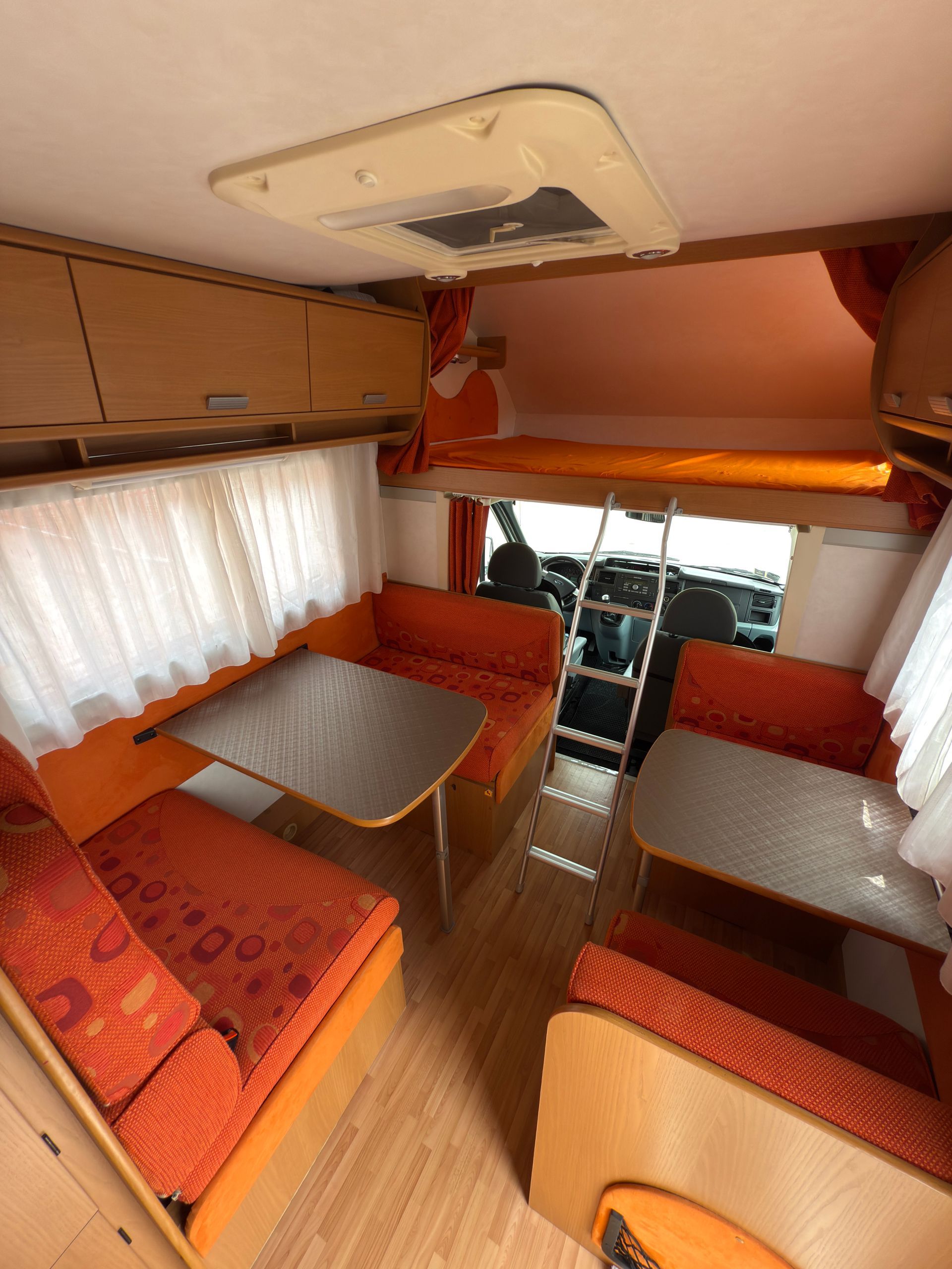 Interno di un camper arancione e beige con due tavoli, posti a sedere, scaletta e zona notte sopraelevata.