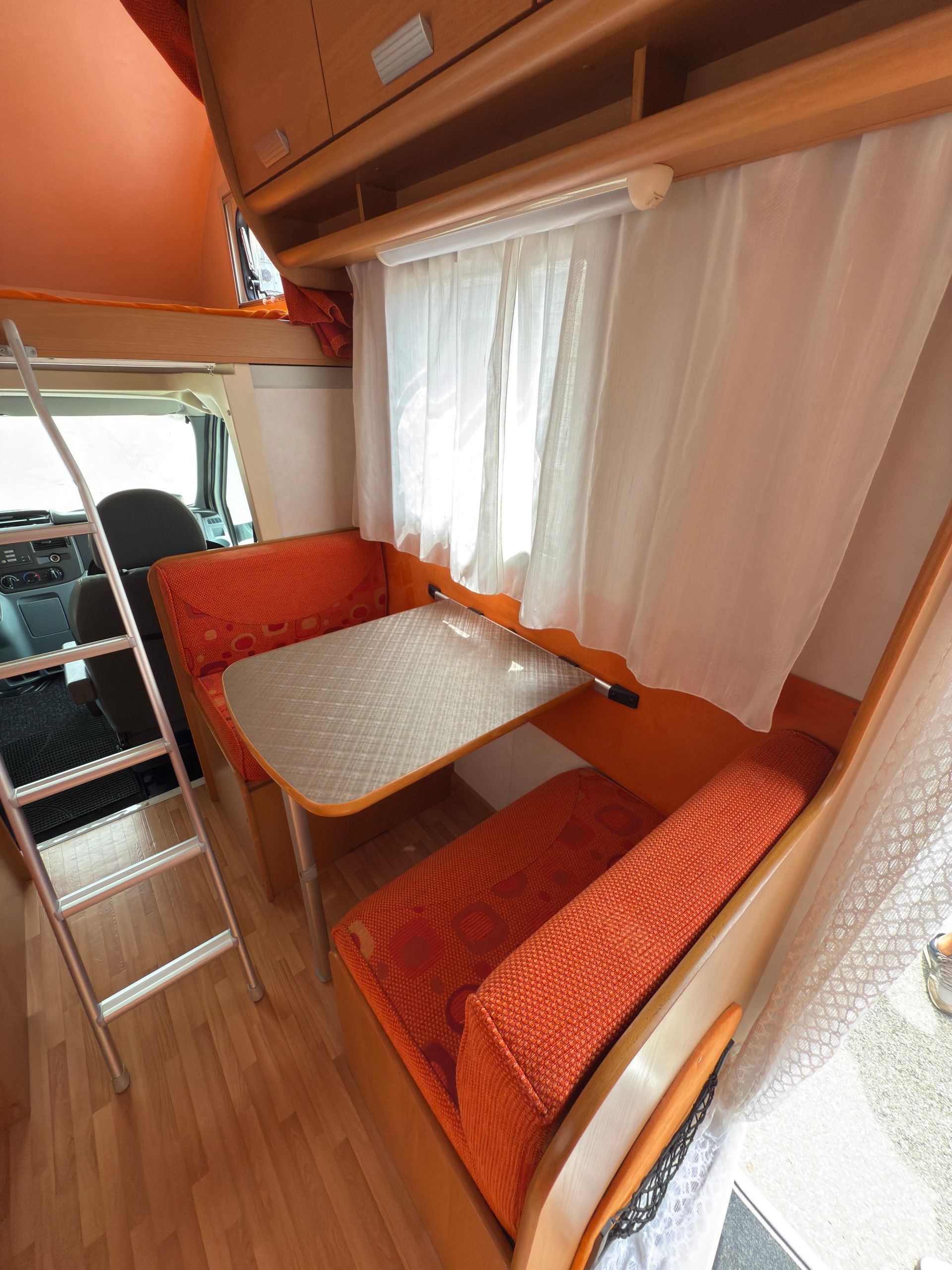 Interno di un camper con sedili arancioni, tavolo, finestra con tende e una scala che porta a un soppalco.
